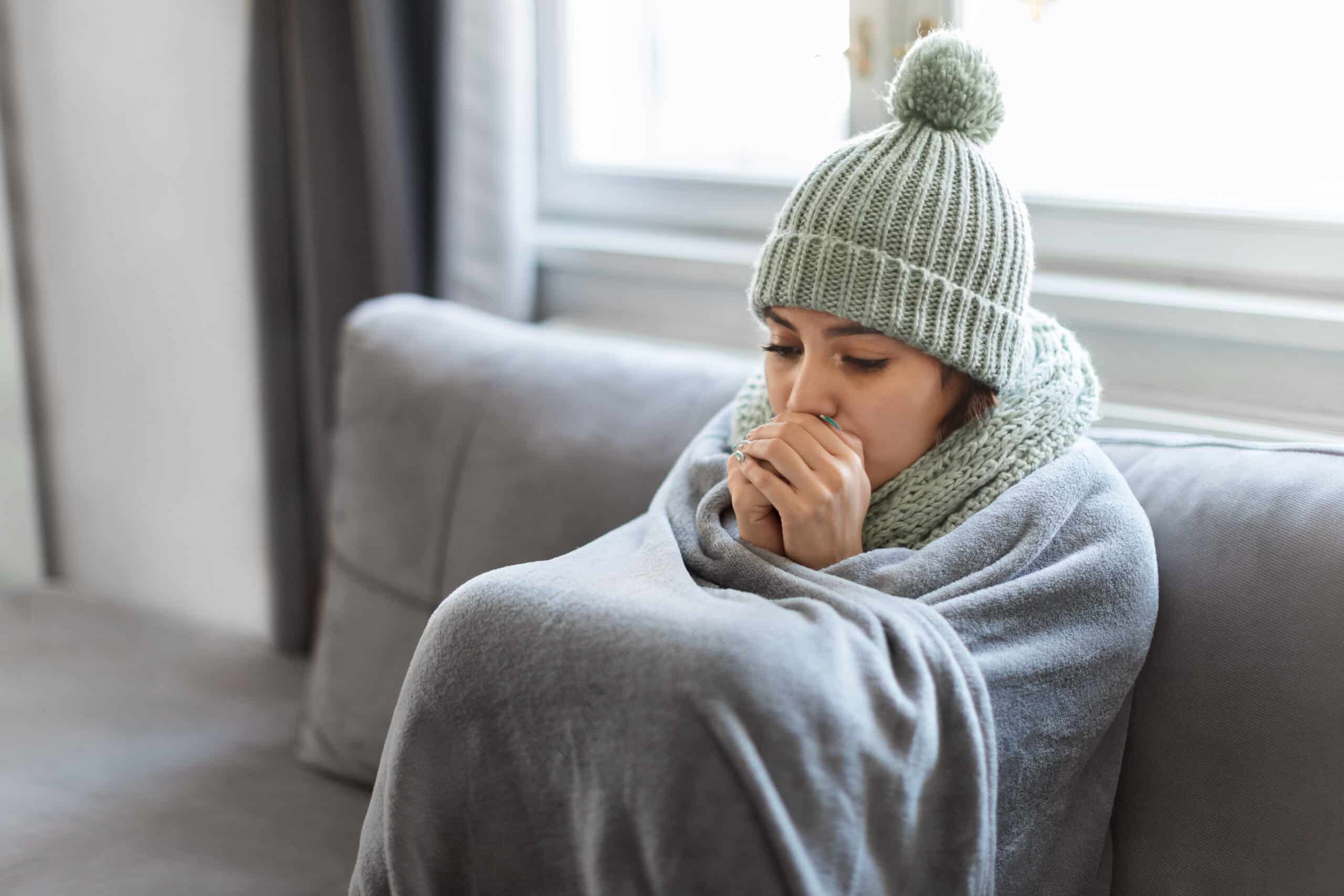 young woman feeling cold at home, sitting on couch covered in blanket